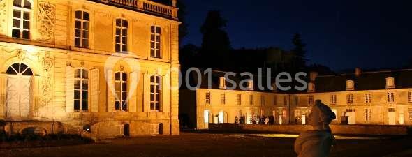 Façade éclairée d'un château de nuit, avec une cour et une statue visible au premier plan.