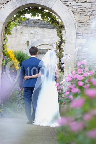 Un couple de mariés de dos traverse une arche en pierre entourée de fleurs dans un jardin.