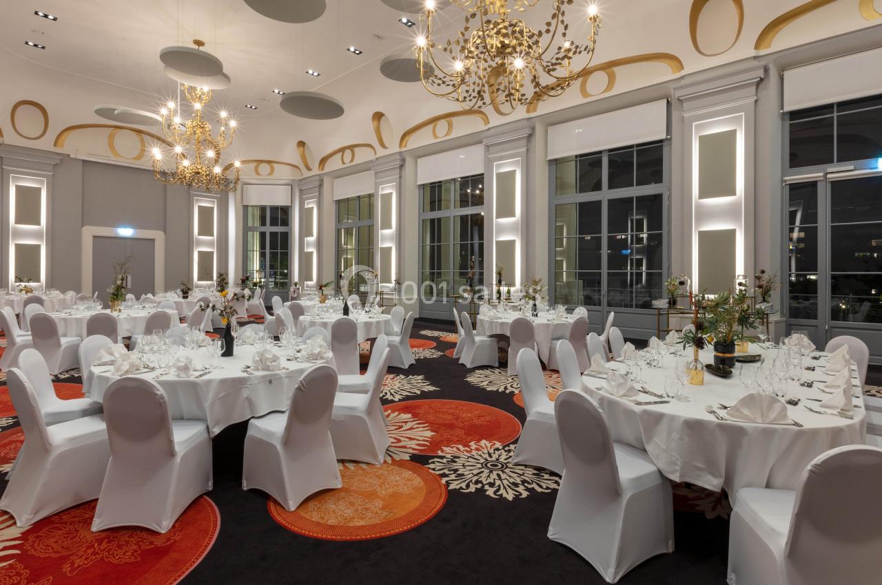 Salle de réception élégante avec tables rondes dressées, chaises blanches et lustres dorés suspendus au plafond.