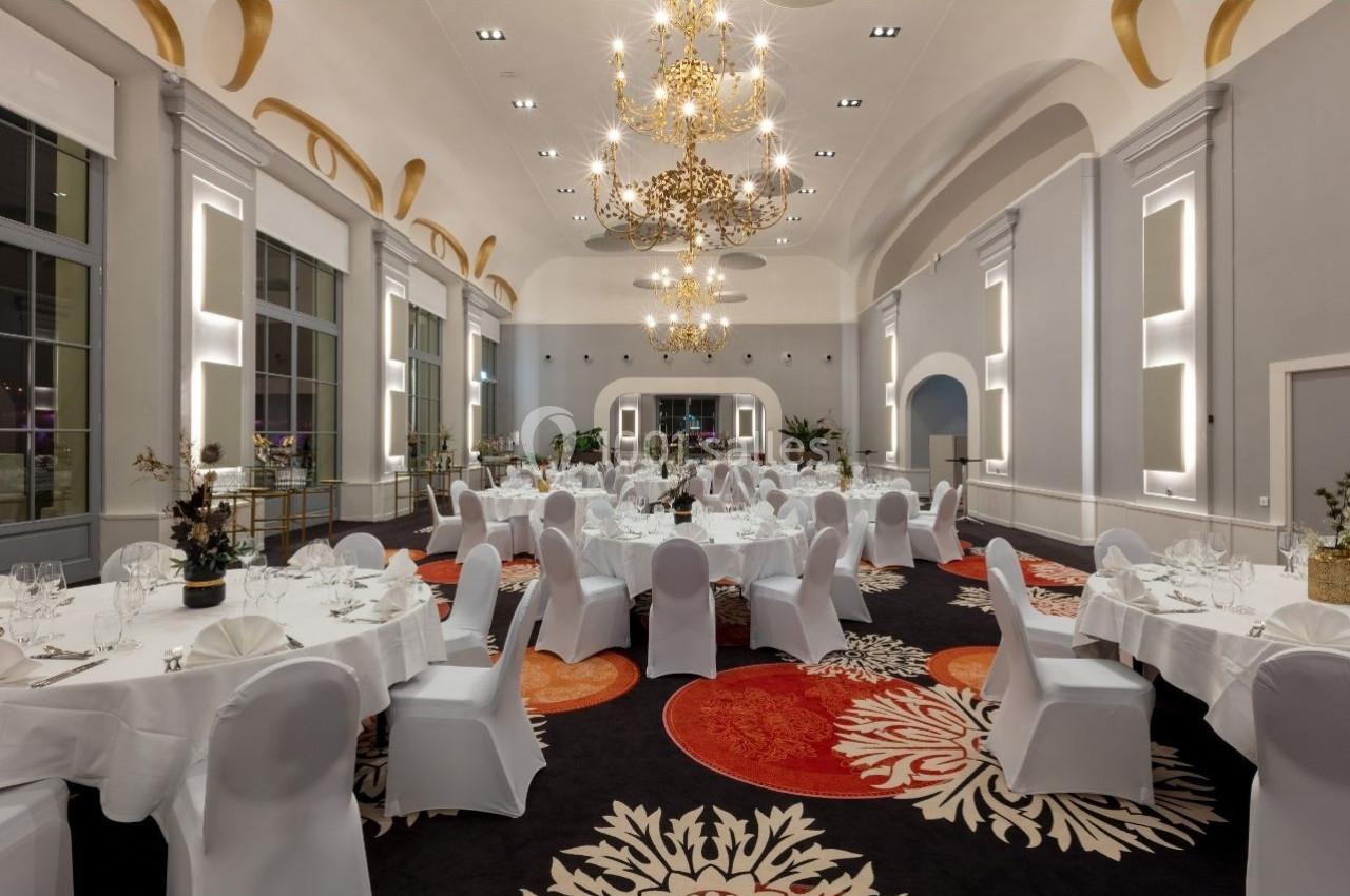 Salle de réception élégante avec tables rondes dressées, chaises blanches et lustres dorés suspendus au plafond.