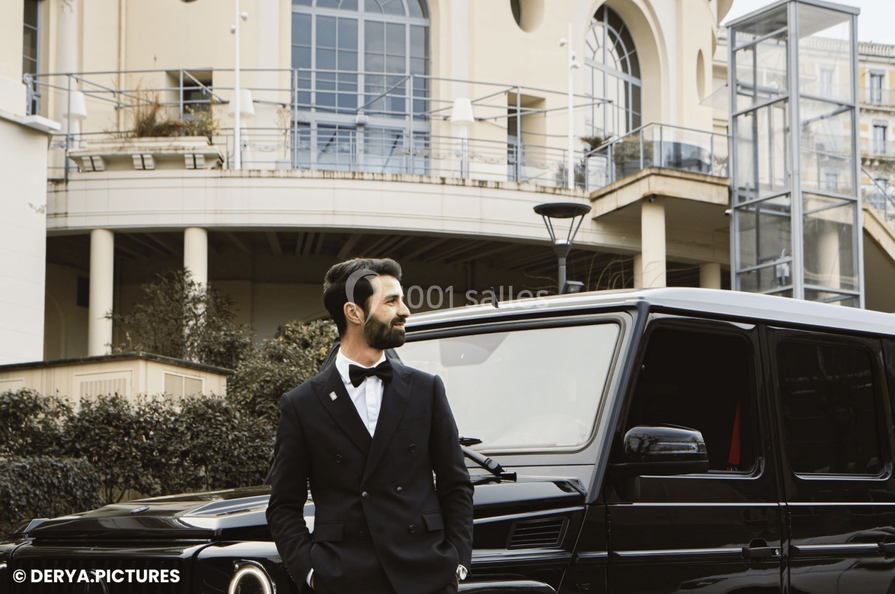 Un homme en costume sombre se tient devant un véhicule noir, avec un bâtiment moderne en arrière-plan.