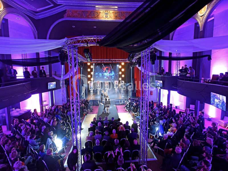 Défilé de mode dans une salle éclairée en violet, avec une scène centrale et un public assis des deux côtés.