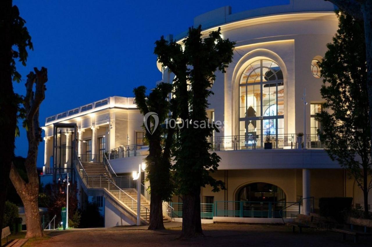 Façade éclairée d'un bâtiment avec de grandes fenêtres, entouré d'arbres, photographié à la tombée de la nuit.