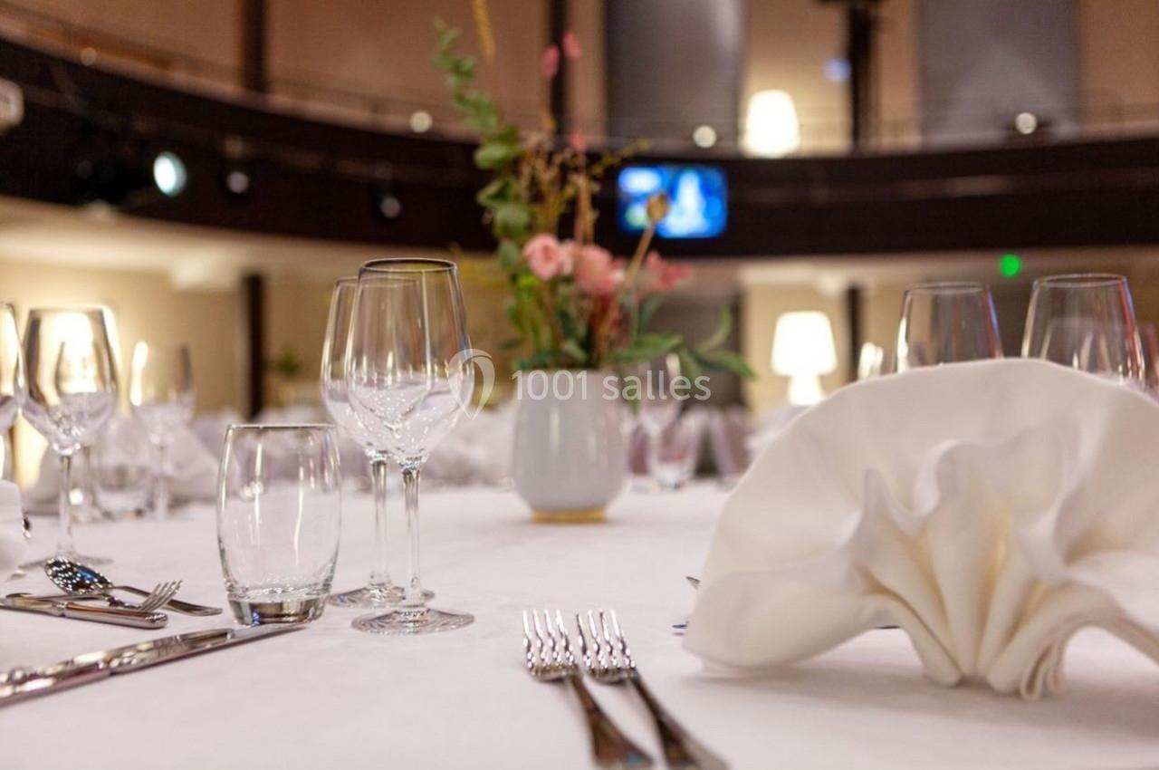Table élégamment dressée avec verres, couverts, serviette pliée et vase de fleurs au centre, dans une salle éclairée.