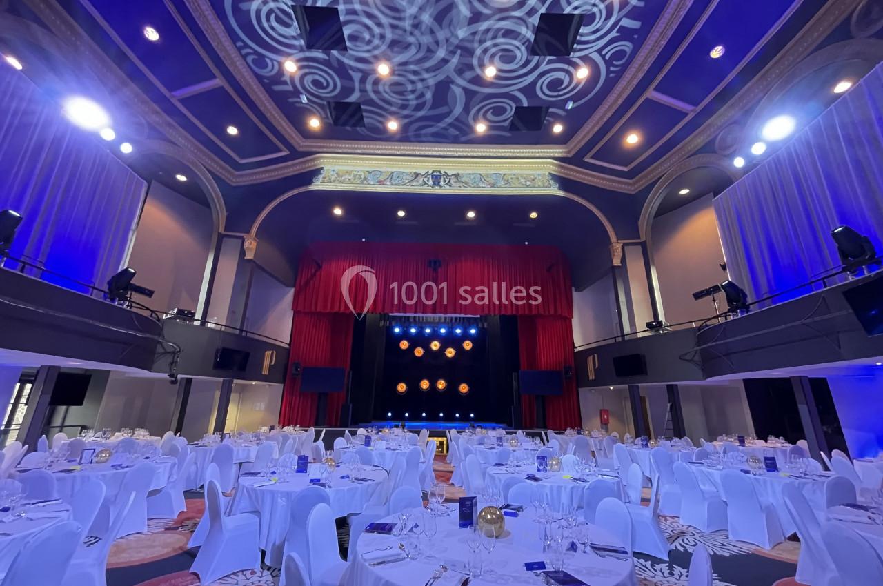 Salle de réception élégante avec tables dressées, éclairage bleu et scène ornée de rideaux rouges.
