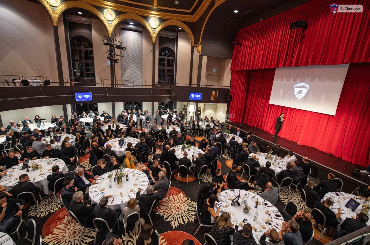 Salle de réception avec des tables rondes occupées par des convives, face à une scène avec un écran de projection.