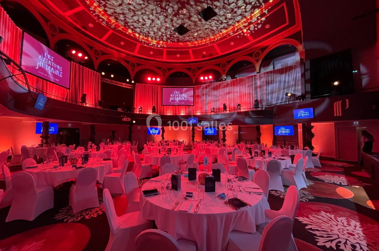 Salle de réception éclairée en rouge avec tables rondes dressées pour un événement, écrans affichant un titre.