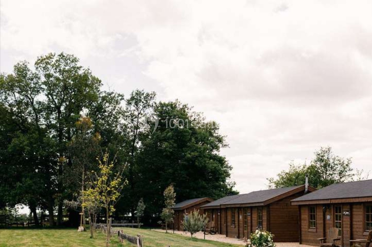 Alignement de petits chalets en bois entourés de verdure, avec des arbres et un ciel nuageux en arrière-plan.