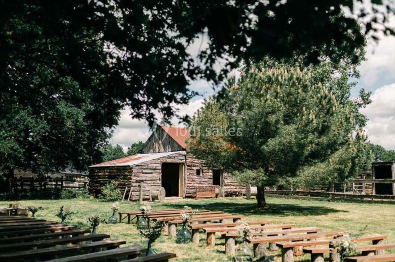 Vue d'une grange en bois entourée d'arbres, avec des bancs disposés en extérieur sur une pelouse.