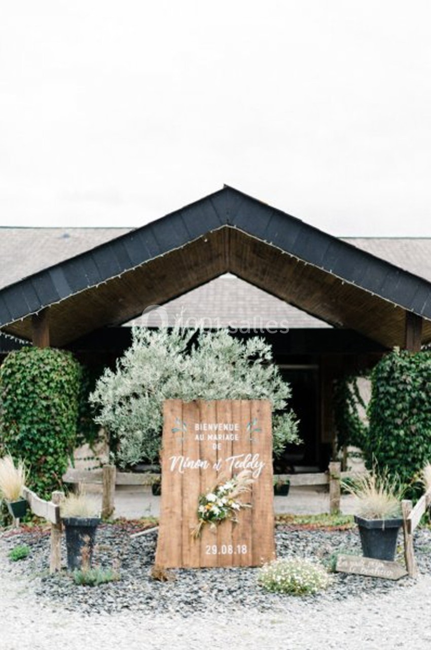 Entrée d'un bâtiment avec un panneau en bois décoré de fleurs, indiquant une date et des prénoms.