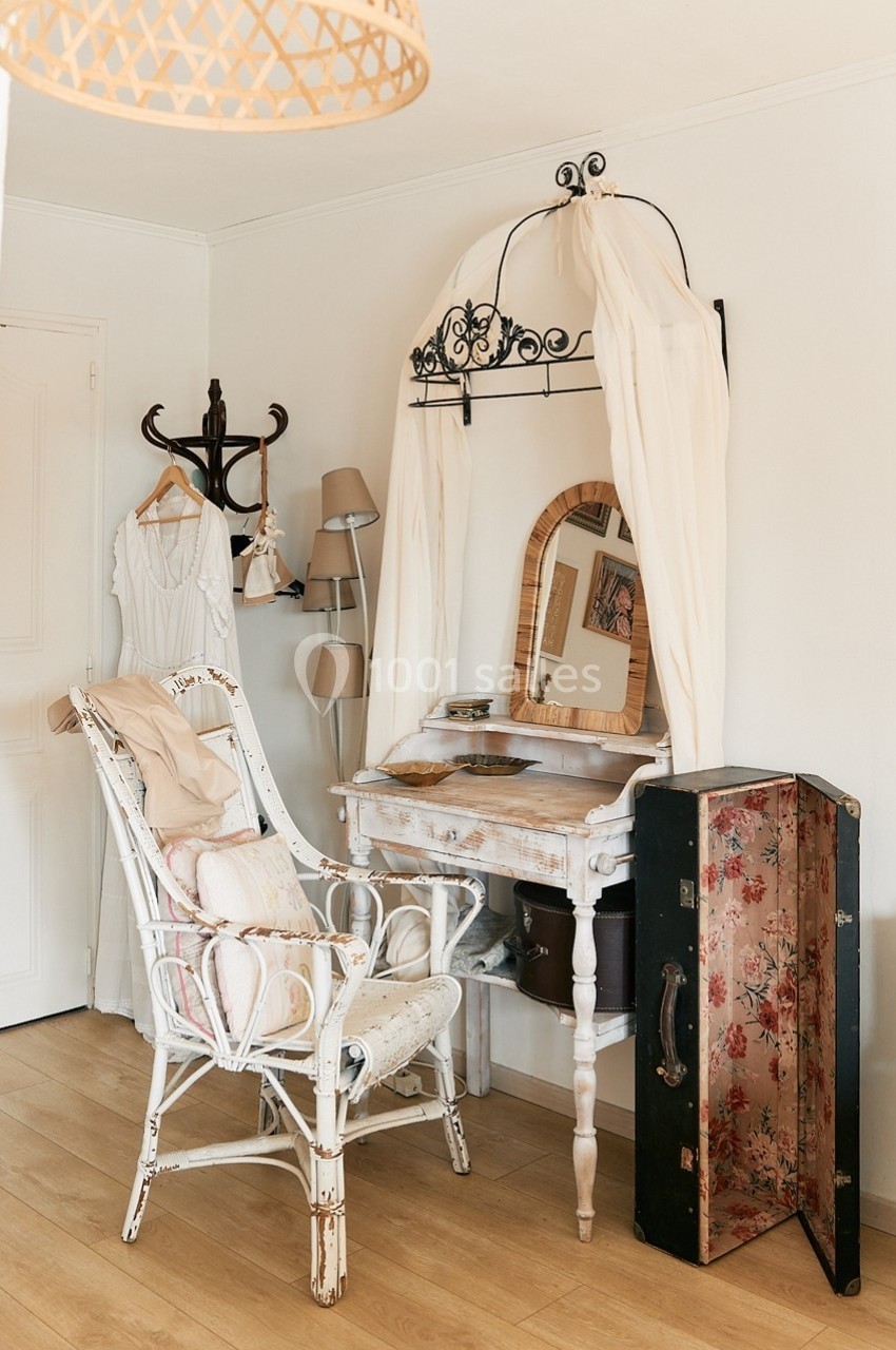 Coiffeuse vintage avec miroir, chaise en osier, et valise ouverte dans une chambre au style rétro et lumineux.
