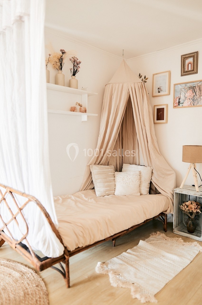 Lit simple avec baldaquin beige, coussins décoratifs, étagères avec vases et cadres accrochés sur un mur blanc.