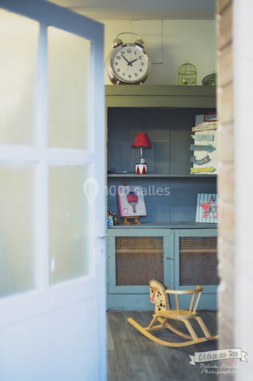 Vue d'une pièce avec une étagère bleue, une horloge, des livres, une lampe rouge et un cheval à bascule en bois.