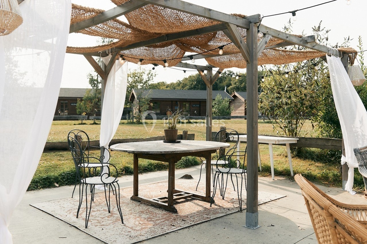 Pergola en bois avec voiles blanches, table ronde et chaises en métal, située dans un jardin verdoyant.