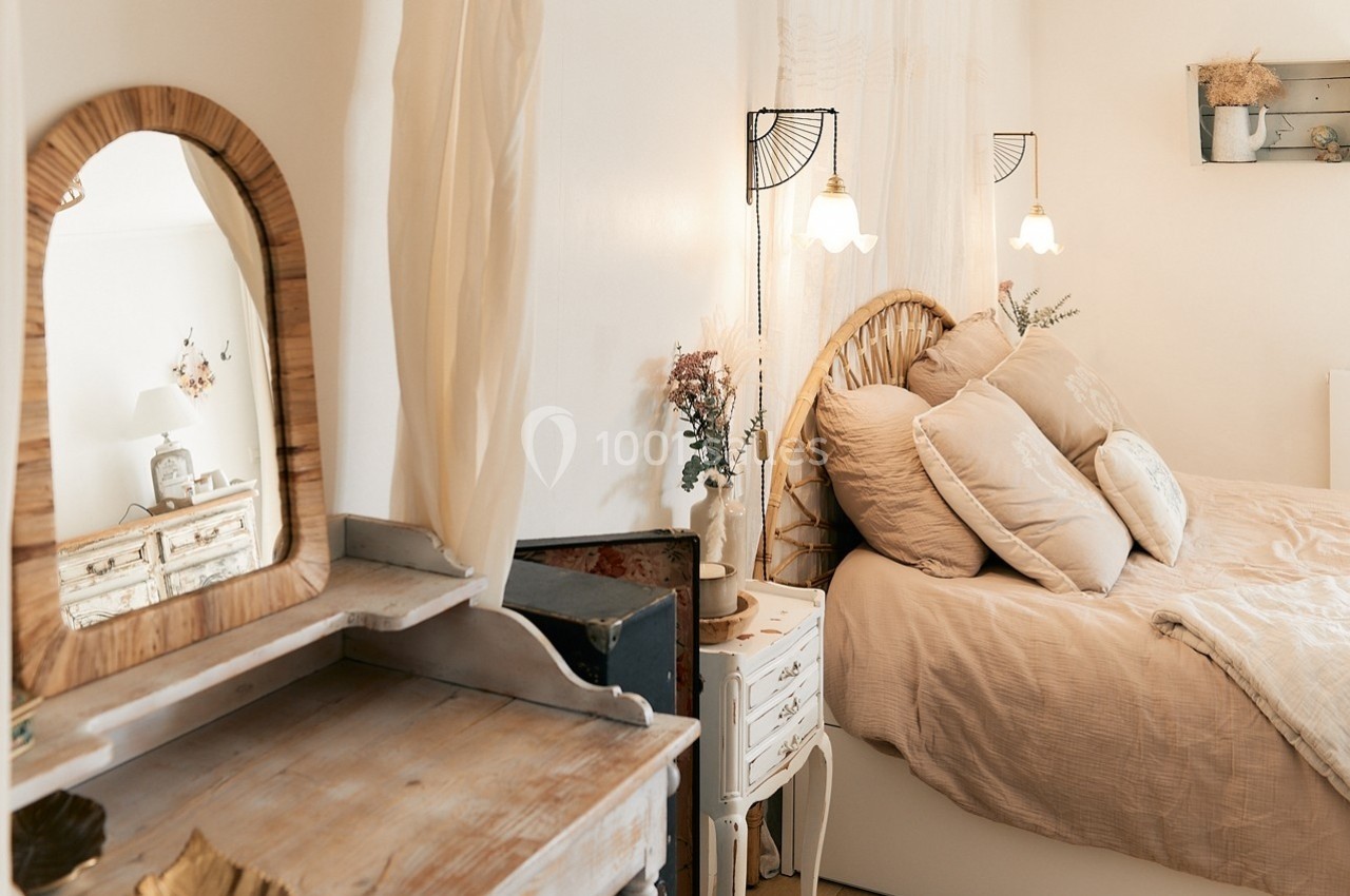 Chambre lumineuse avec lit en rotin, coussins beiges, coiffeuse en bois et lampes suspendues au style vintage.