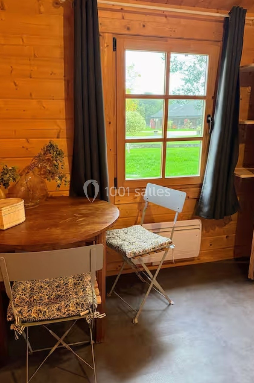 Intérieur d'un chalet en bois avec une table ronde, deux chaises pliantes et une fenêtre donnant sur un jardin.