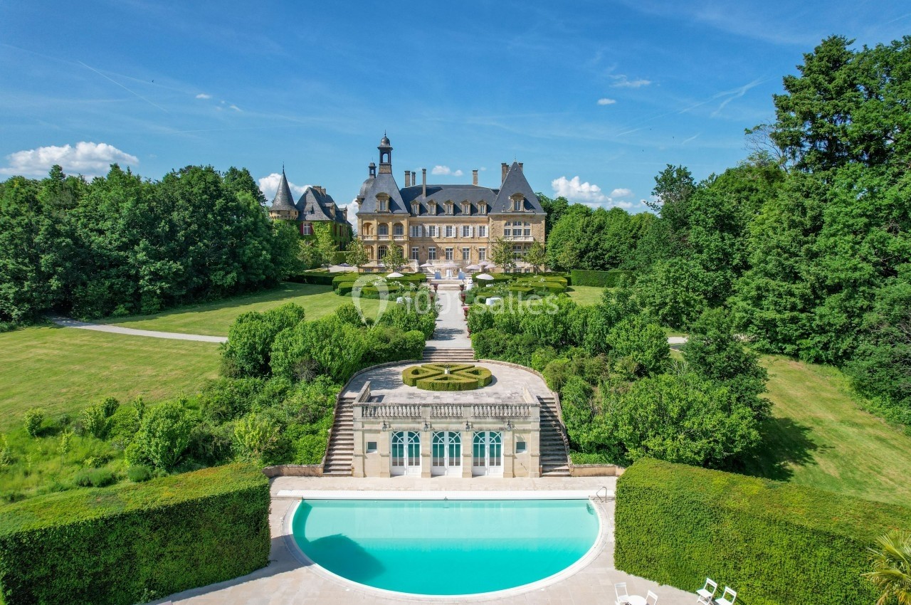 Vue aérienne d'un château entouré de jardins paysagers, avec une piscine au premier plan et des arbres en arrière-plan.