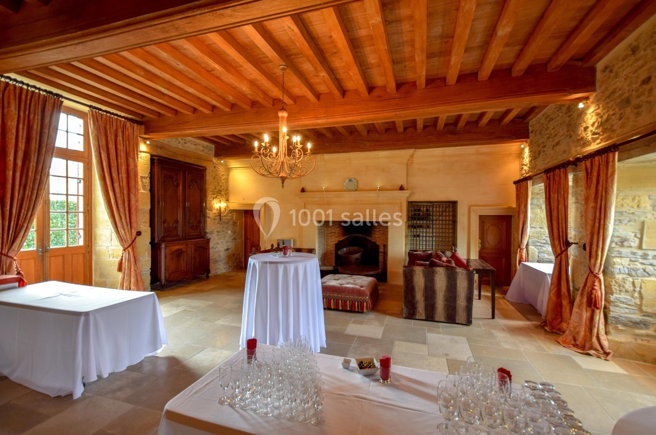 Salle de réception avec poutres apparentes, cheminée en pierre, tables dressées et grandes fenêtres avec rideaux.
