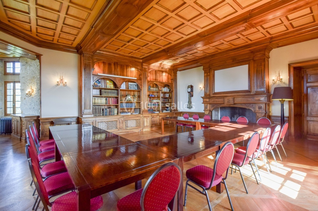 Salle de réunion élégante avec table en bois, chaises rouges, bibliothèque garnie et cheminée dans un décor boisé.