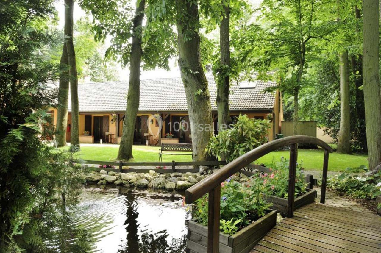 Maison entourée d'arbres avec un jardin, un étang et un petit pont en bois au premier plan.