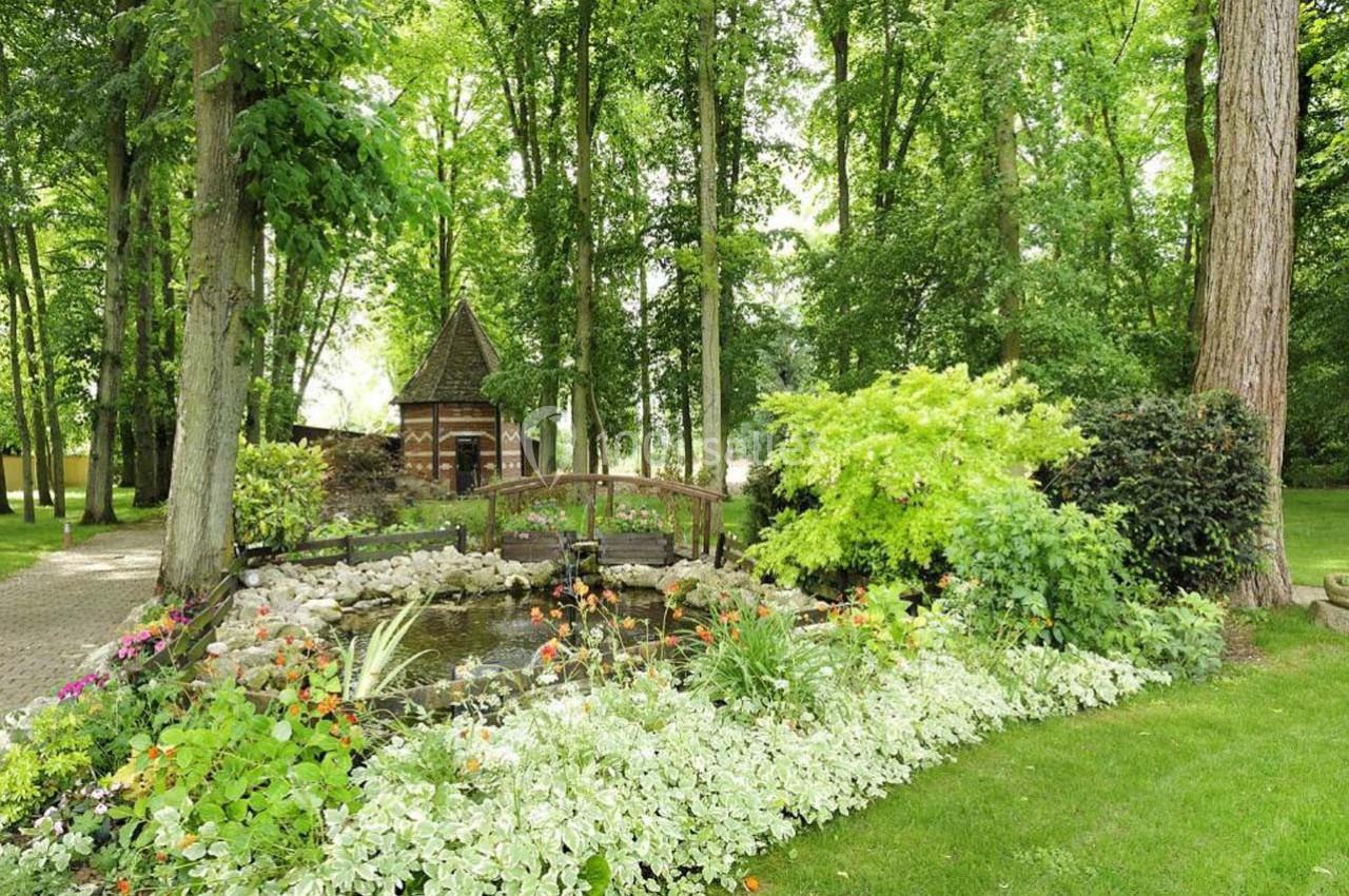 Petit pont en bois surplombant un étang entouré de fleurs et de verdure dans un jardin boisé.