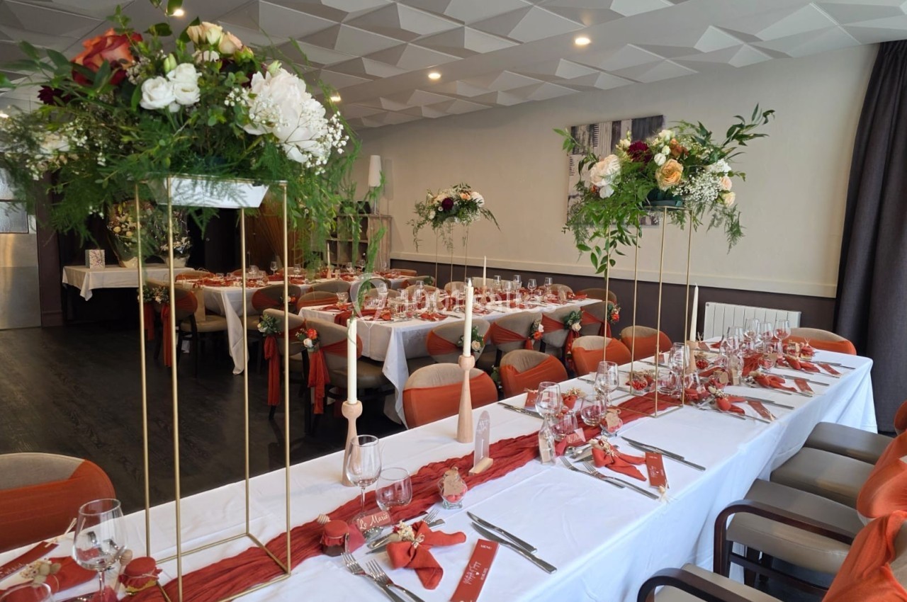 Salle de réception décorée avec des tables dressées, nappes blanches, chemins de table rouges et compositions florales.