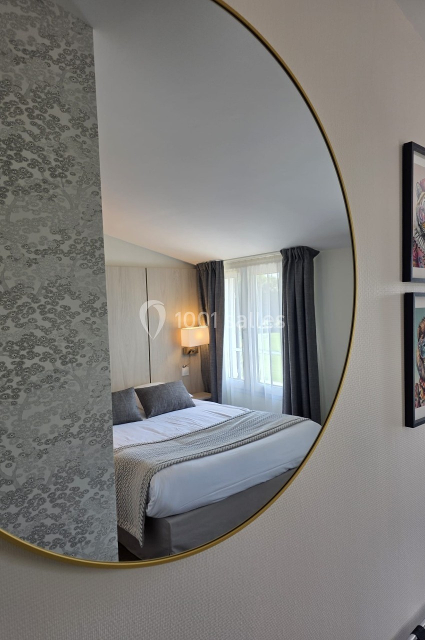 Reflet d'une chambre lumineuse avec lit double, rideaux gris et tableaux colorés, vu dans un miroir rond doré.