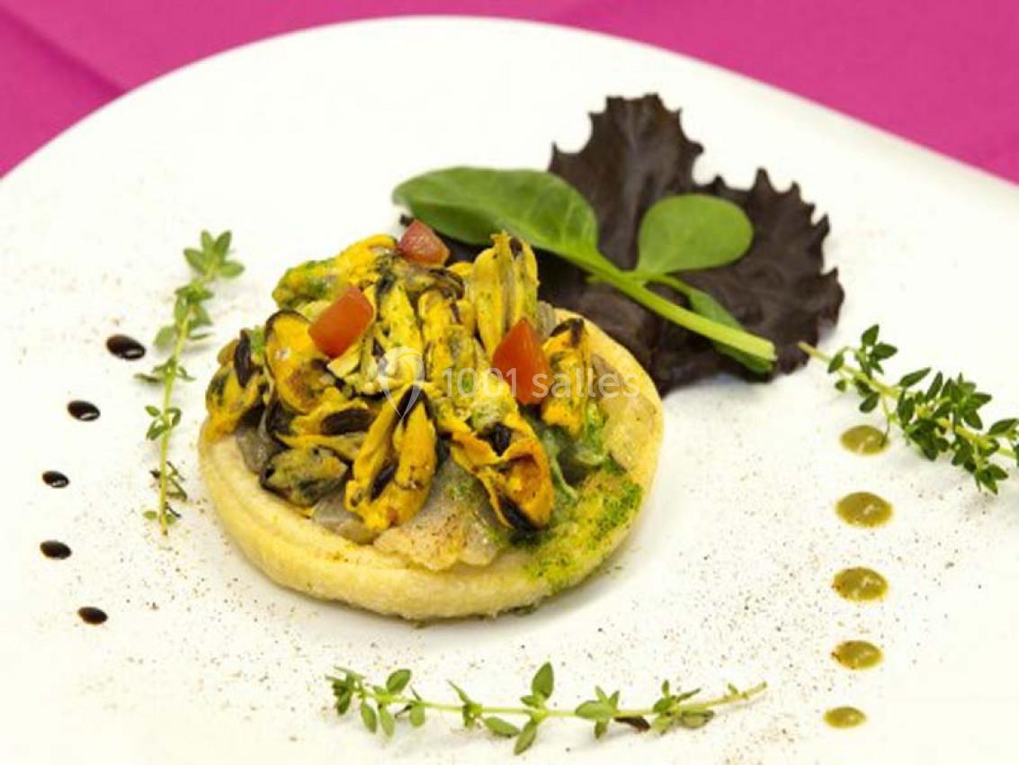 Tartelette garnie de moules, sauce verte et dés de tomate, servie avec des feuilles de salade sur une assiette blanche.