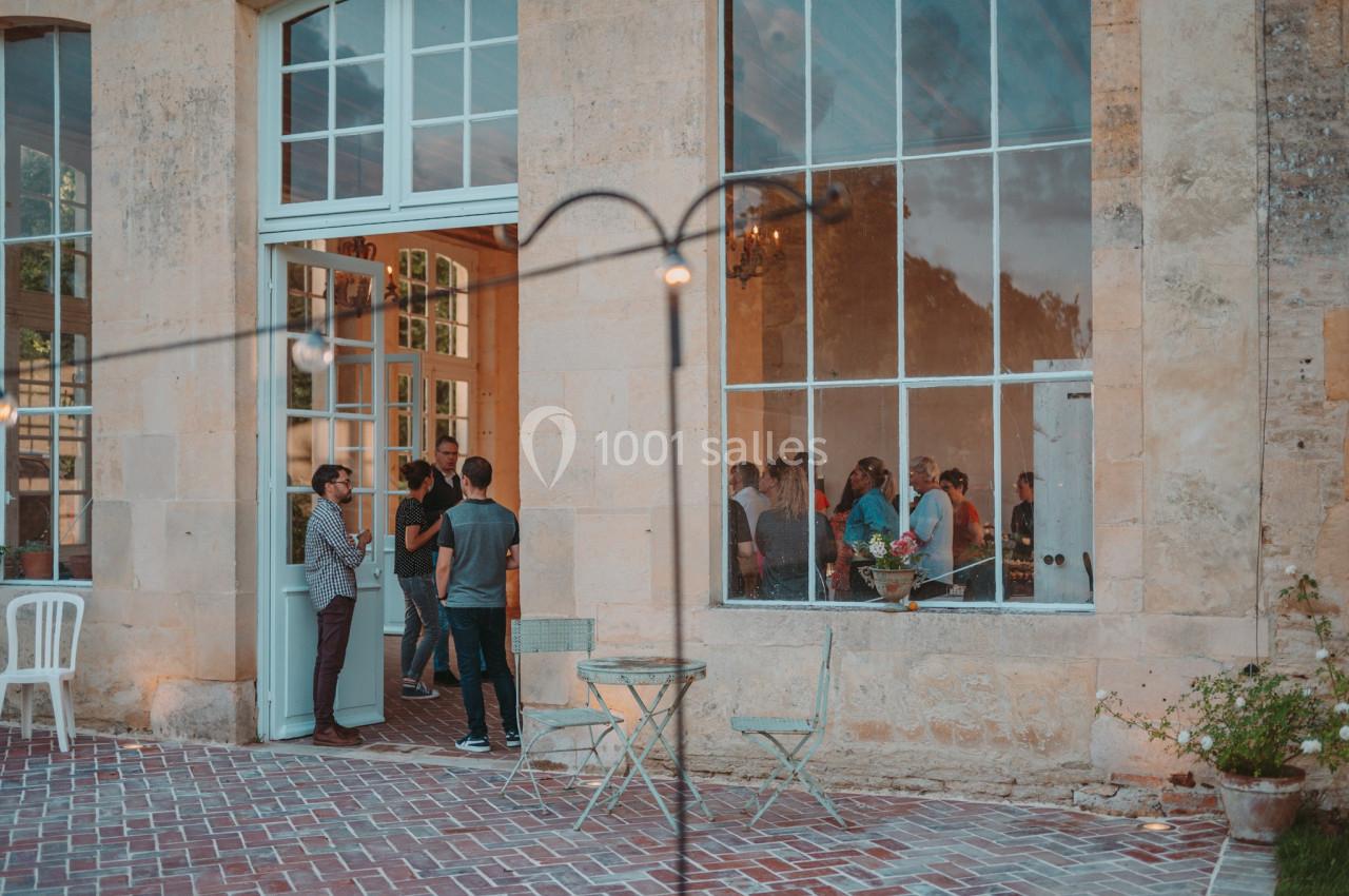 Groupe de personnes discutant à l'entrée d'un bâtiment en pierre avec de grandes fenêtres, tables et chaises en extérieur.