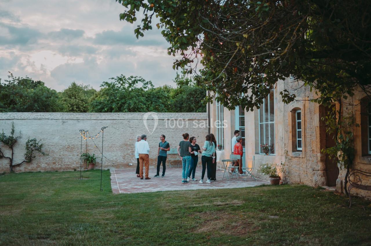 Groupe de personnes discutant dans une cour pavée près d'une maison en pierre, entourée de verdure.