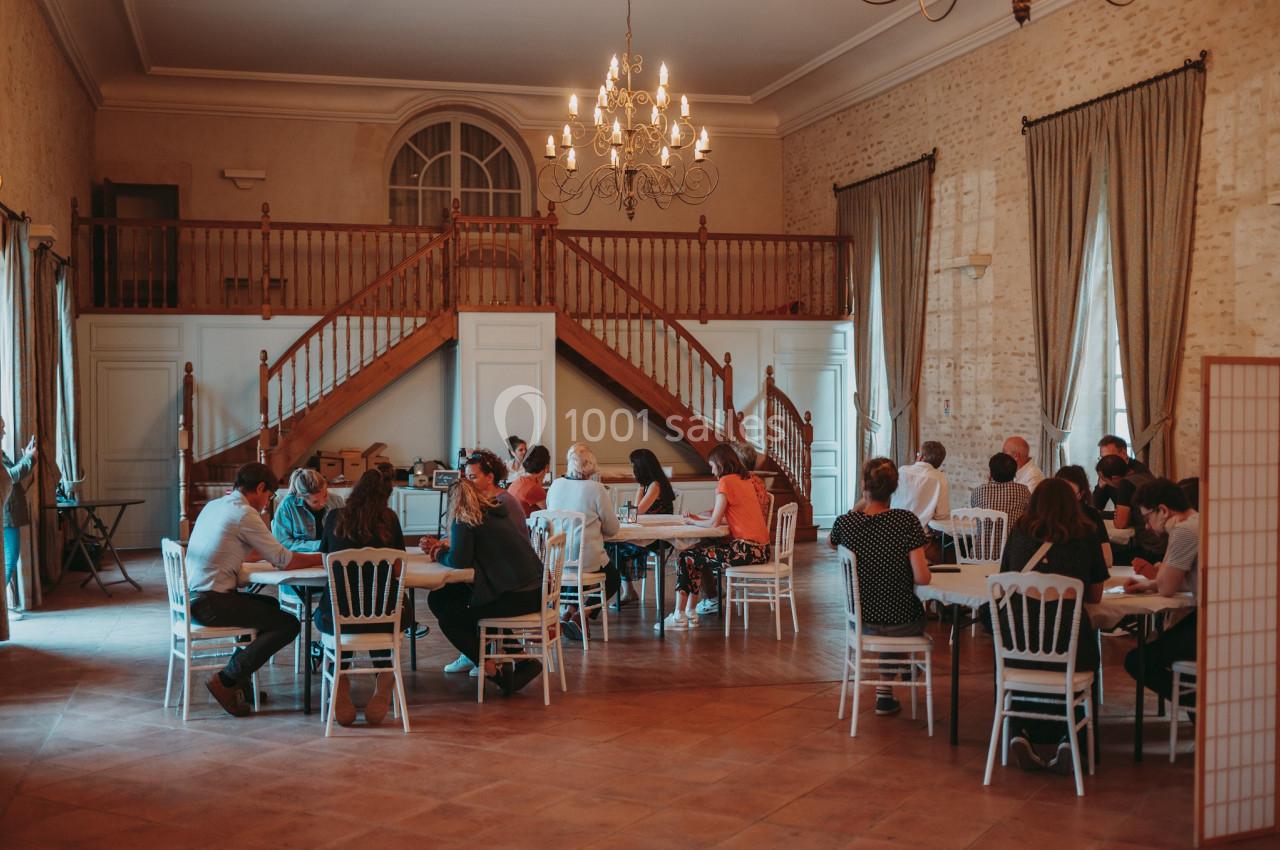 Personnes assises en petits groupes autour de tables dans une grande salle avec un escalier en bois et un lustre.