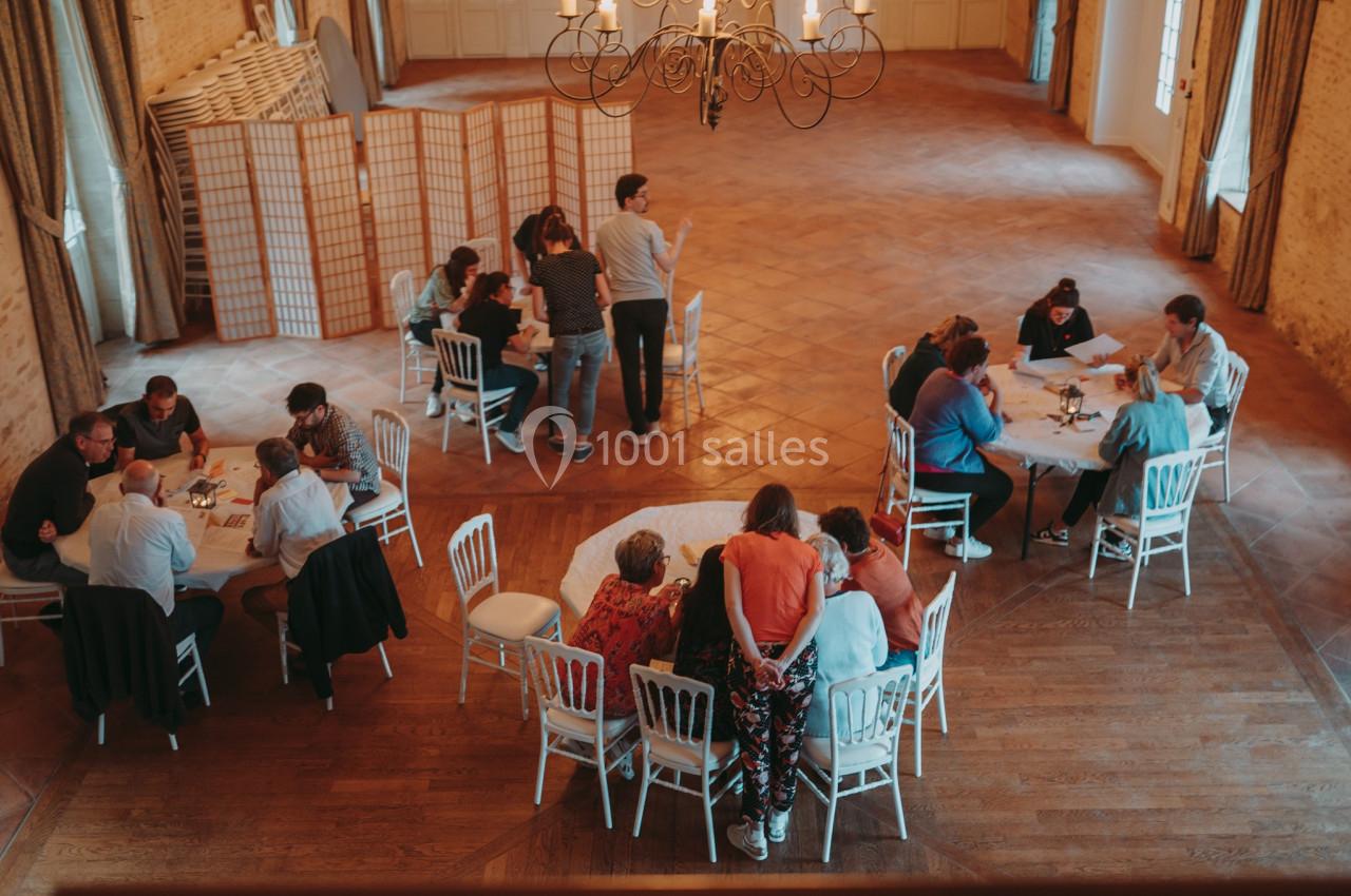 Des groupes de personnes discutent autour de tables rondes dans une grande salle avec parquet et cloison décorative.