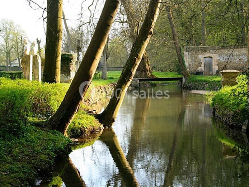 Rivière bordée d'arbres inclinés, avec une petite bâtisse en pierre et des statues dans un cadre verdoyant.