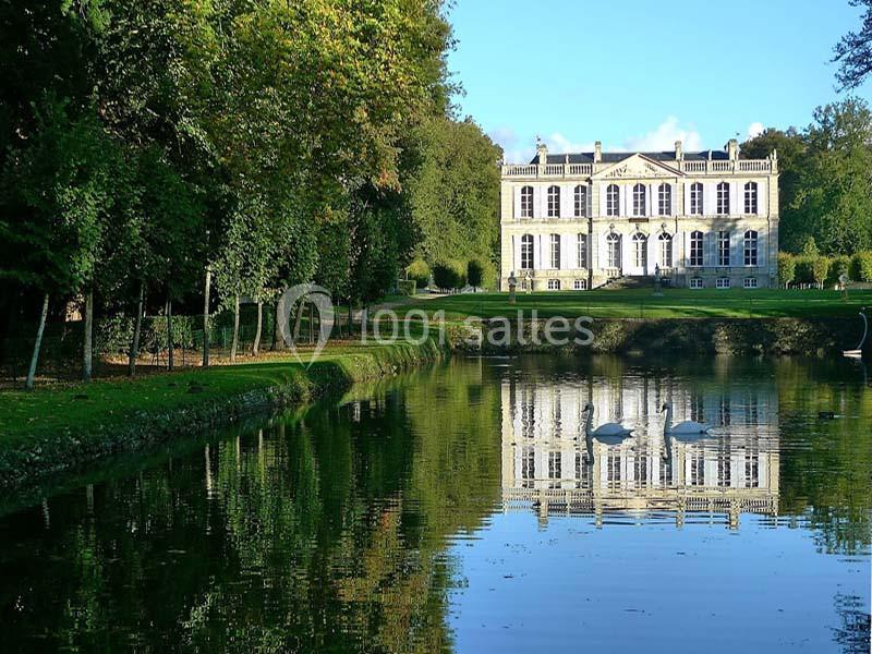 Façade d'un château blanc entouré d'arbres, avec son reflet visible dans un étang au premier plan.