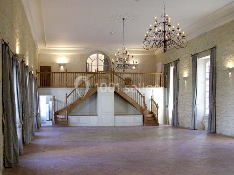 Salle spacieuse avec murs en pierre, lustres suspendus, mezzanine en bois et grandes fenêtres encadrées de rideaux.