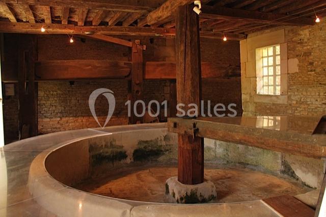 Intérieur d'un ancien moulin avec une meule en pierre centrale, des murs en pierre et un plafond en bois.