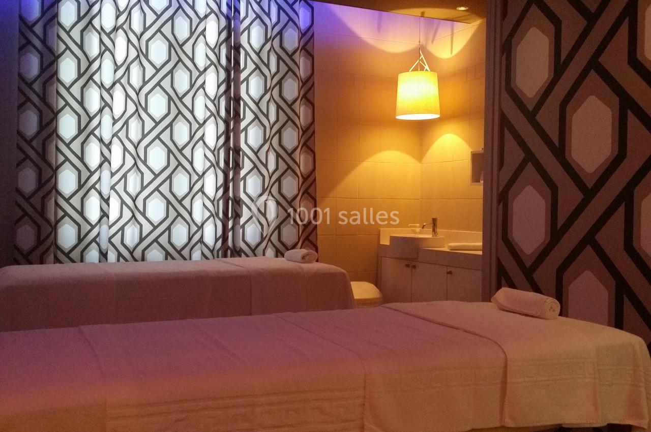 Salle de massage avec deux tables recouvertes de draps blancs, éclairage tamisé et motifs géométriques sur les murs.