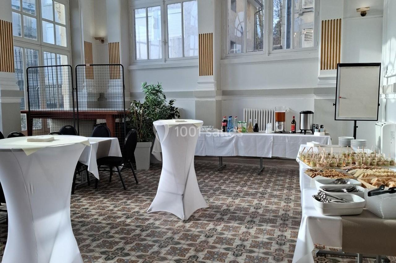 Salle lumineuse avec grandes fenêtres, tables hautes habillées de nappes blanches et buffet de boissons et snacks.