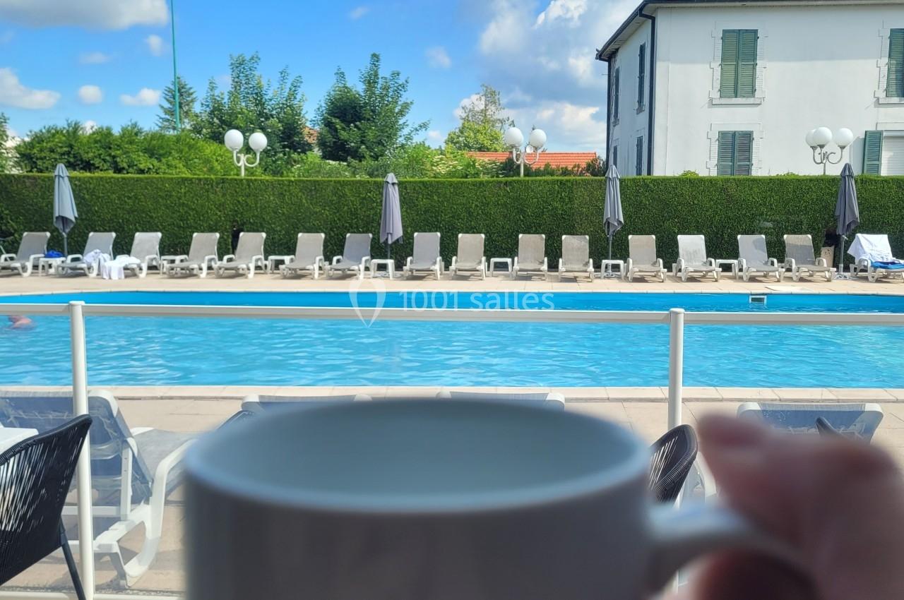 Une tasse tenue devant une piscine extérieure entourée de chaises longues et d'un bâtiment sous un ciel bleu.