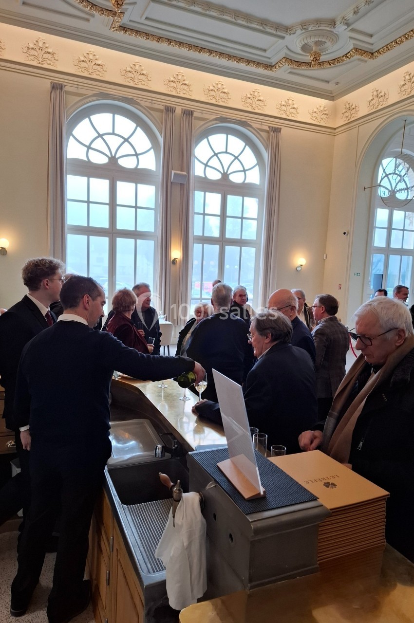 Un groupe de personnes discute près d'un comptoir dans une salle lumineuse avec de grandes fenêtres et un plafond orné.