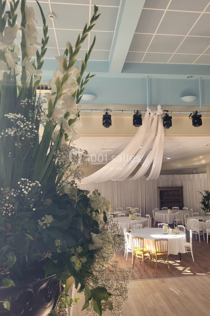 Salle de réception lumineuse avec tables rondes dressées, chaises blanches et grande composition florale au premier plan.