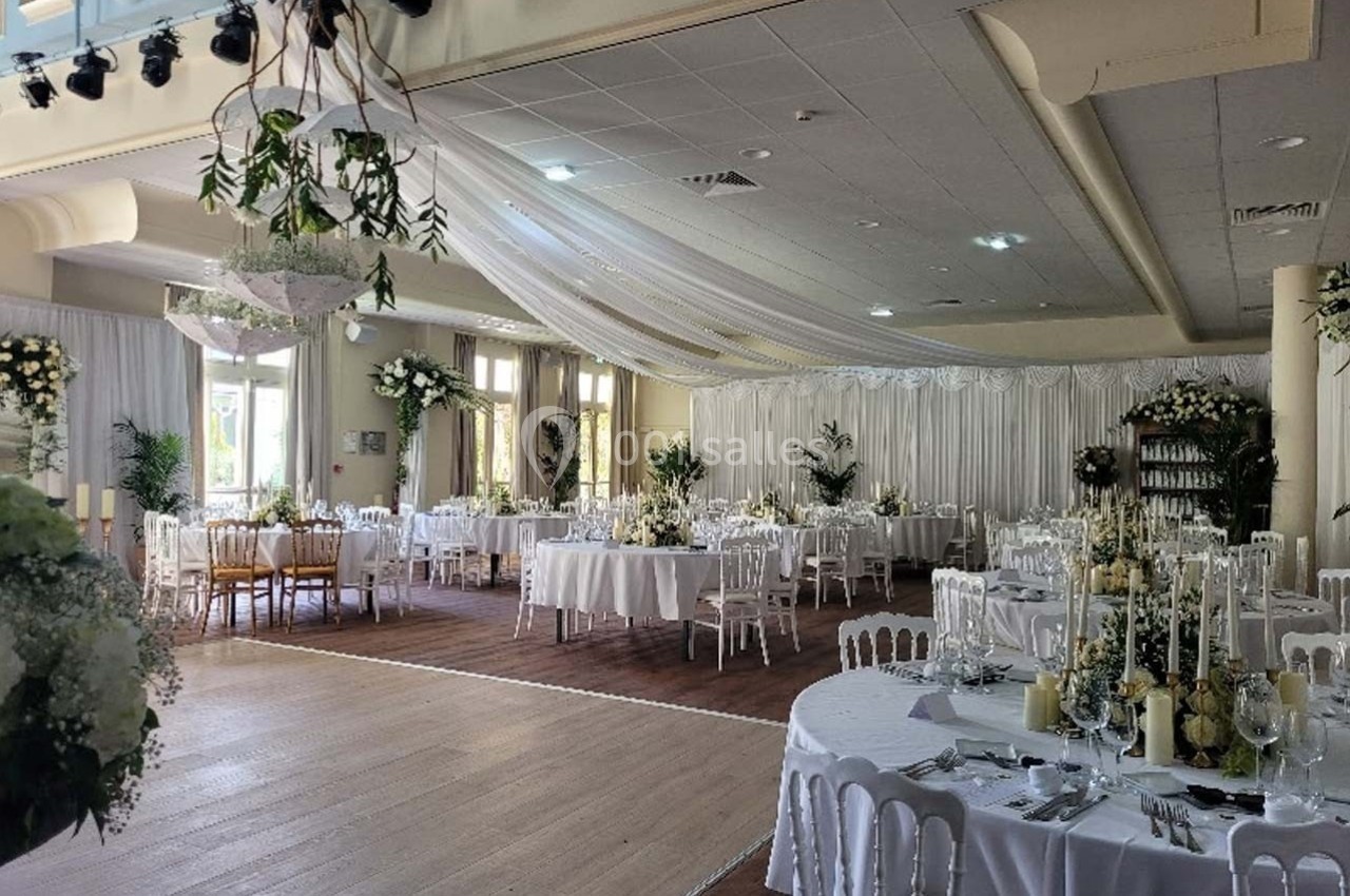 Salle de réception élégamment décorée avec des tables rondes, nappes blanches, fleurs et éclairage naturel.