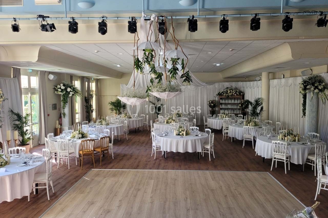 Salle de réception décorée pour un mariage, avec tables rondes, chaises blanches et suspensions végétales.