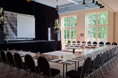 Salle de conférence lumineuse avec tables disposées en U, écran de projection et grandes fenêtres donnant sur l'extérieur.