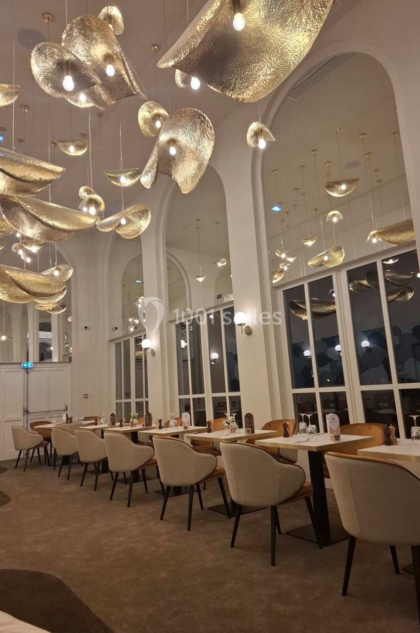 Salle de restaurant élégante avec des tables dressées, des chaises beige et des luminaires dorés suspendus au plafond.