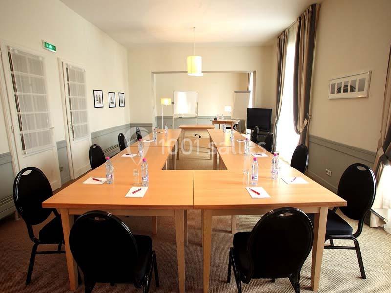 Salle de réunion lumineuse avec tables en U, chaises noires, bouteilles d'eau et bloc-notes disposés sur les tables.