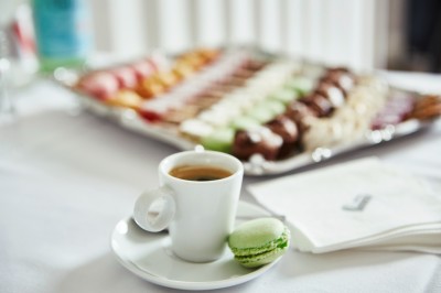 Tasse de café sur une table avec un macaron vert et un plateau de desserts flous en arrière-plan.