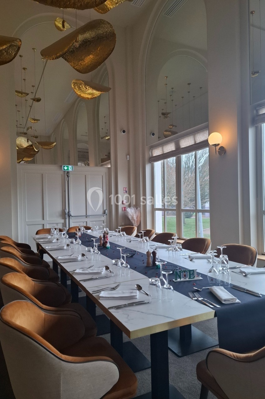 Salle de restaurant élégante avec une grande table dressée, chaises en velours et décorations dorées suspendues.