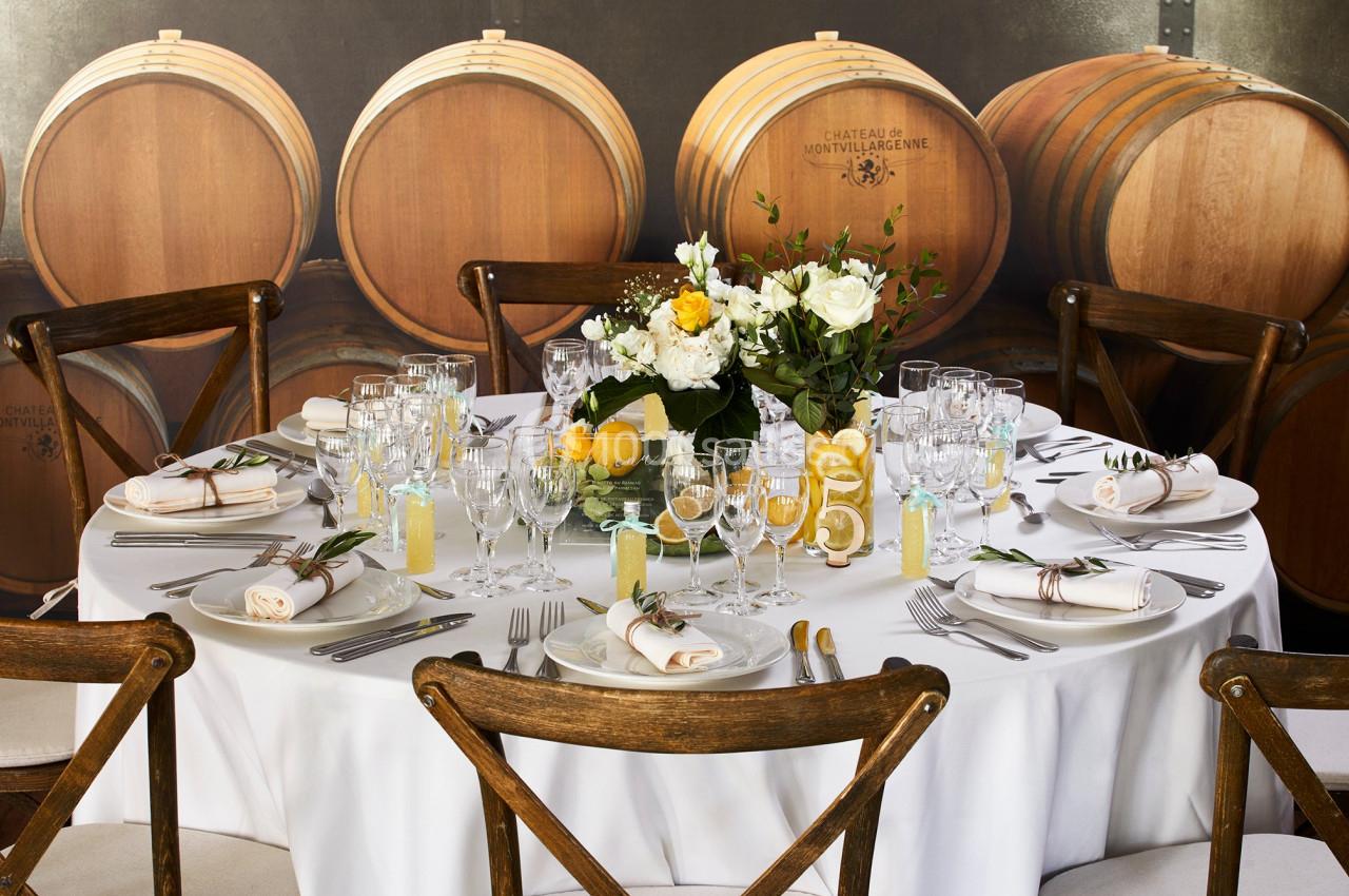 Table ronde élégamment dressée avec vaisselle raffinée, décor floral et tonneaux en bois en arrière-plan.