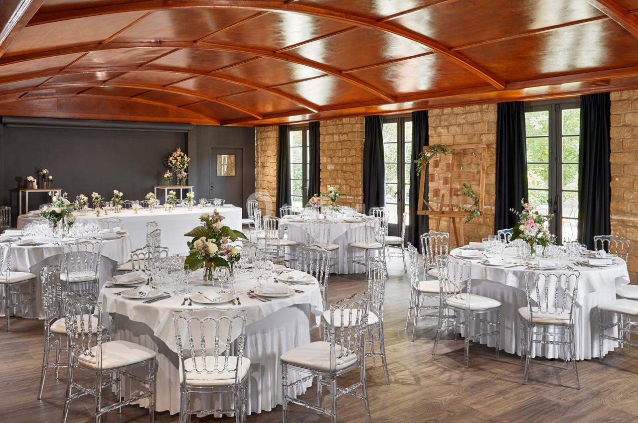 Salle de réception élégante avec tables rondes dressées, chaises transparentes et décoration florale, éclairée par des…